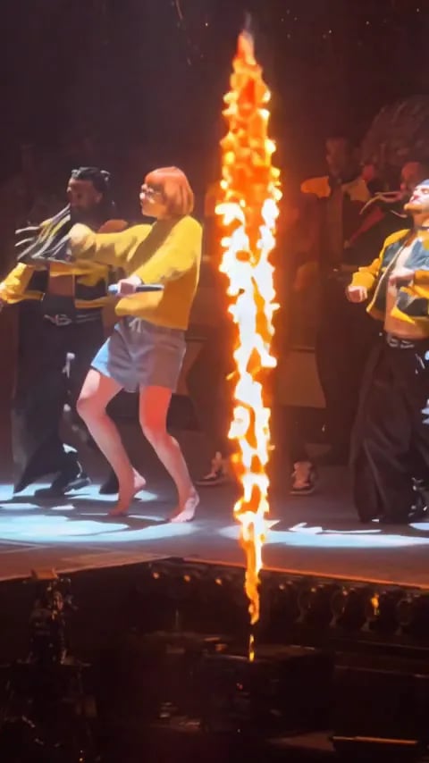 A woman with a red bob, glasses and a mustard sweater leads the backup dancers on a concert stage next to a flame jet